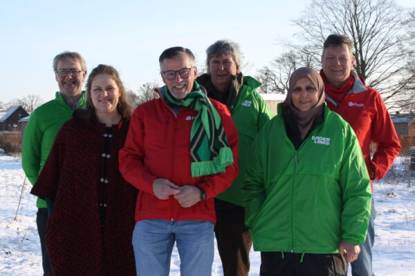 Vier mannen en twee vrouwen met rode en groene jasjes aan staan in een groepje lachend in de sneeuw. Op de achtergrond wat bomen.